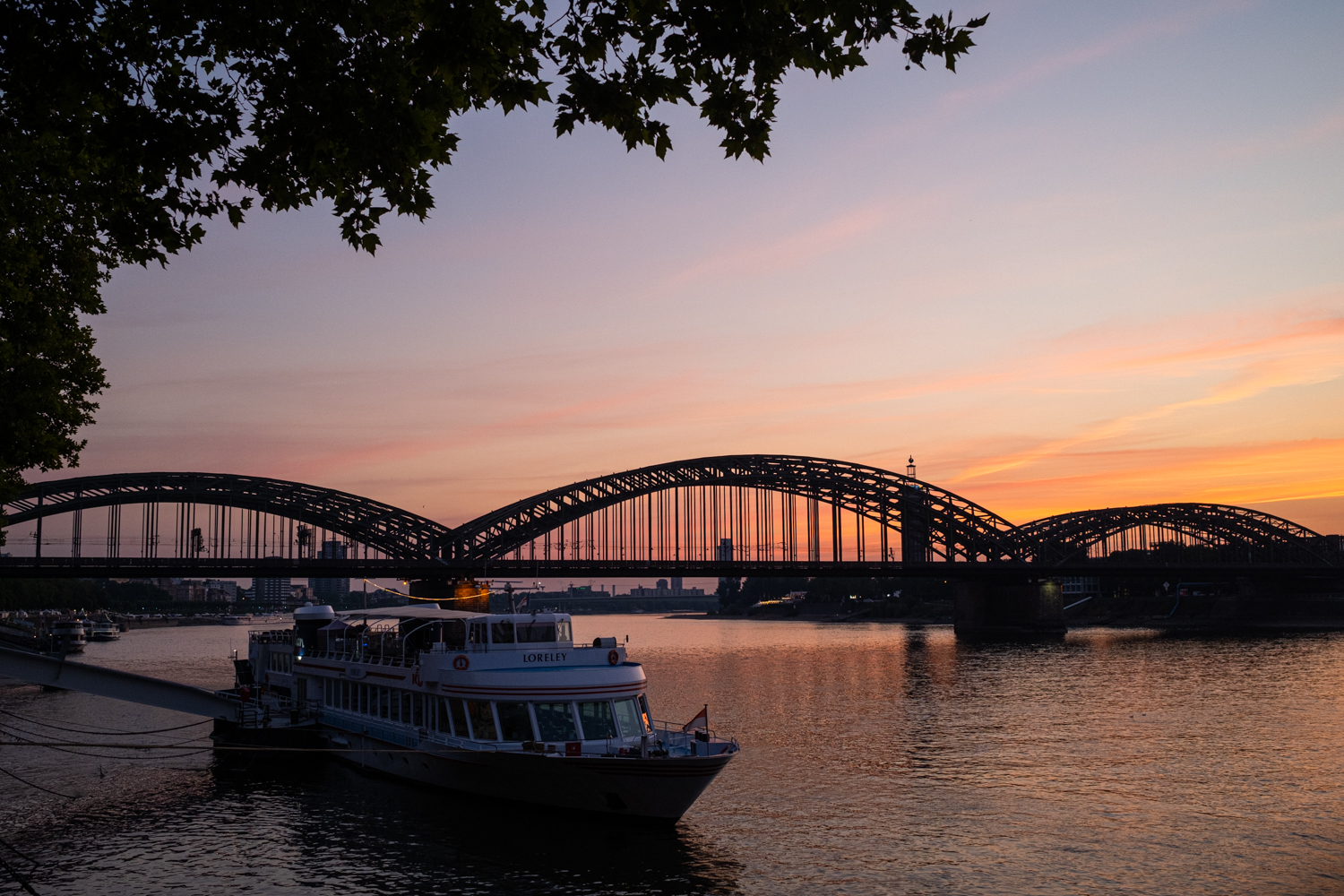 Blick auf die Hohenzollernbrücke in Köln. Im Vordergrund ist ein Ausflugsschiff zu sehen. Das Foto entstand kurz vor Sonnenaufgang.